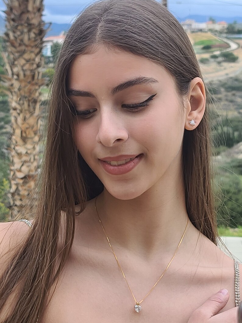 Pu&ograve; includere: Una persona giovane con lunghi capelli castani indossa una collana a catena dorata con un piccolo ciondolo a forma di cuore. Il ciondolo ha una pietra trasparente al centro.