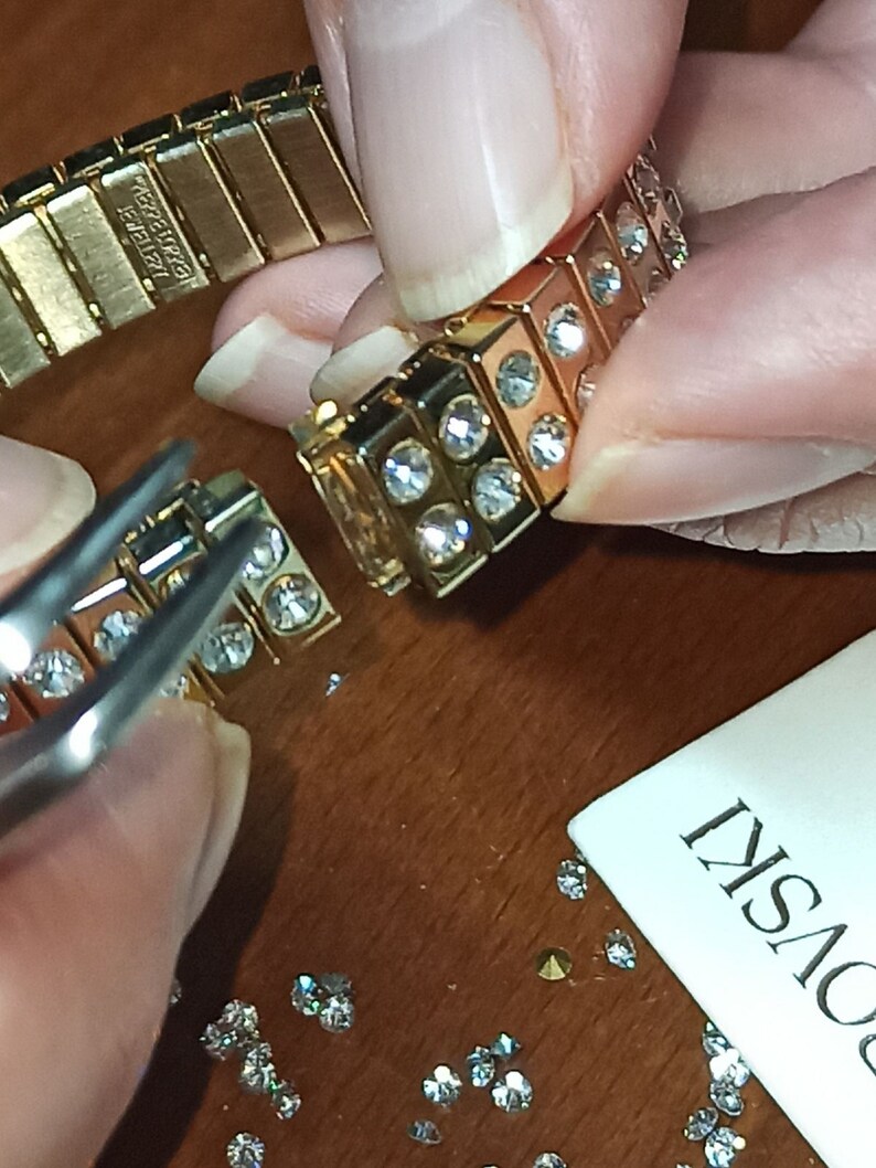 Puede incluir: Un brazalete dorado con una fila de cristales transparentes. El brazalete se est&aacute; ensamblando con pinzas y cristales sueltos.