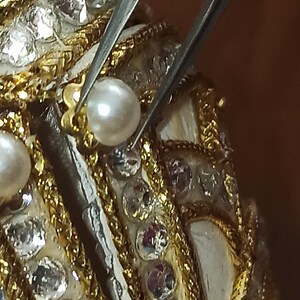 May include: Close-up of a white egg-shaped object decorated with gold trim and small, clear rhinestones. A pair of tweezers is holding a white pearl in place on the object.