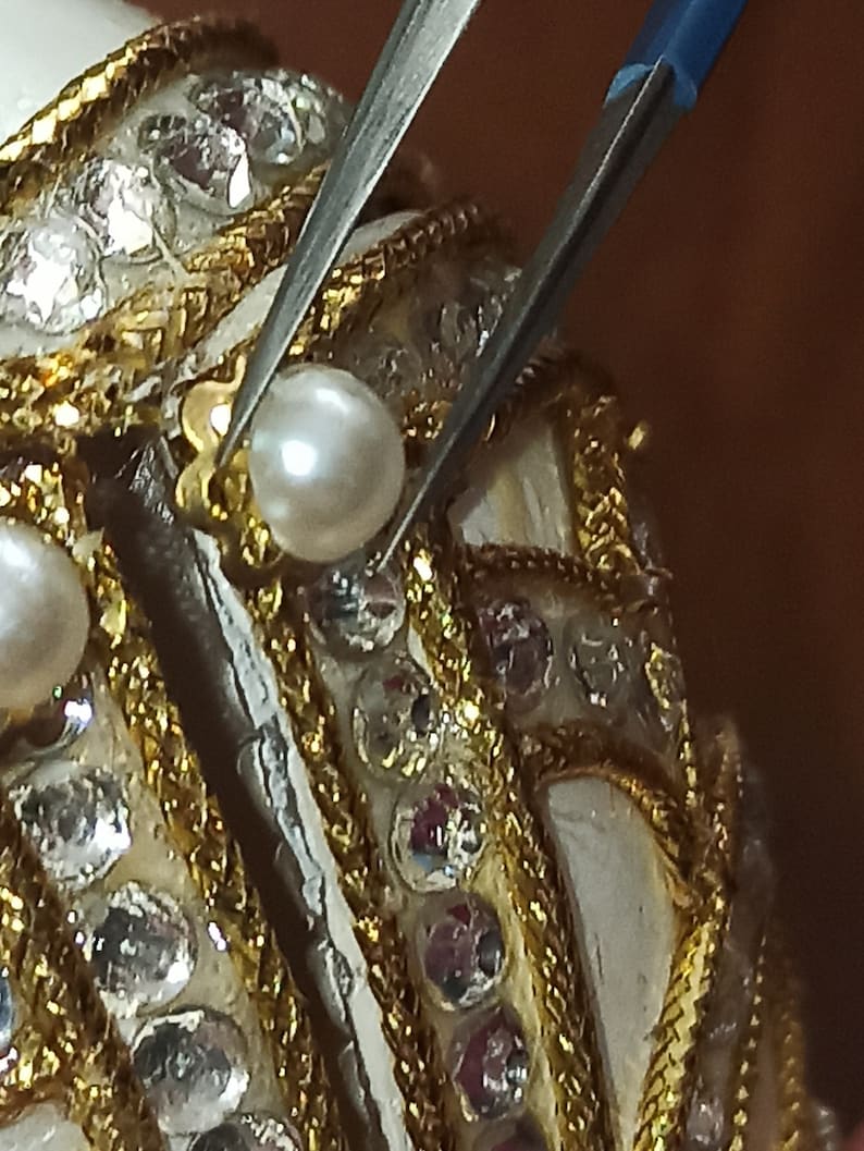 May include: Close-up of a white egg-shaped object decorated with gold trim and small, clear rhinestones. A pair of tweezers is holding a white pearl in place on the object.