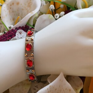 May include: A silver and gold bracelet with red gemstones. The bracelet is made of metal links and has a magnetic clasp.