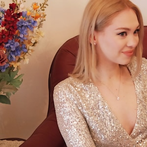 Pu&ograve; includere: Una donna con i capelli biondi indossa un abito di paillettes argentate con scollatura profonda. &Egrave; seduta su una poltrona rossa e sorride.