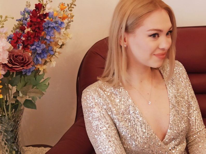 Pu&ograve; includere: Una donna con i capelli biondi indossa un abito di paillettes argentate con scollatura profonda. &Egrave; seduta su una poltrona rossa e sorride.