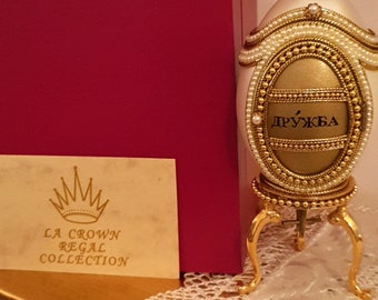 May include: A white egg-shaped decorative object with gold accents and a pearl-like top. The object is on a gold stand and has the word "Дружба" written on it. The object is part of the La Crown Regal Collection.