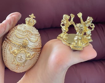 May include: A gold-tone decorative egg-shaped trinket box with a crown on top and a white and gold design. The box is open and has a small gold-tone crown inside. The box is sitting on a hand, next to a gold-tone decorative stand with three lions.