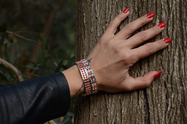 May include: A woman's wrist adorned with two silver bracelets featuring rows of red and clear rhinestones. The bracelets are stacked on top of each other.