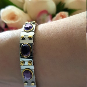Puede incluir: Un brazalete de plata y oro con piedras preciosas púrpuras. El brazalete está hecho de piezas de metal enlazadas y tiene un cierre magnético.