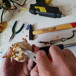 May include: A person is using pliers to attach a gold-colored crown to a white and gold decorative egg. The egg is sitting on a white surface. There are various tools and craft supplies scattered around the egg, including a hammer, a glue gun, and a pair of tweezers.