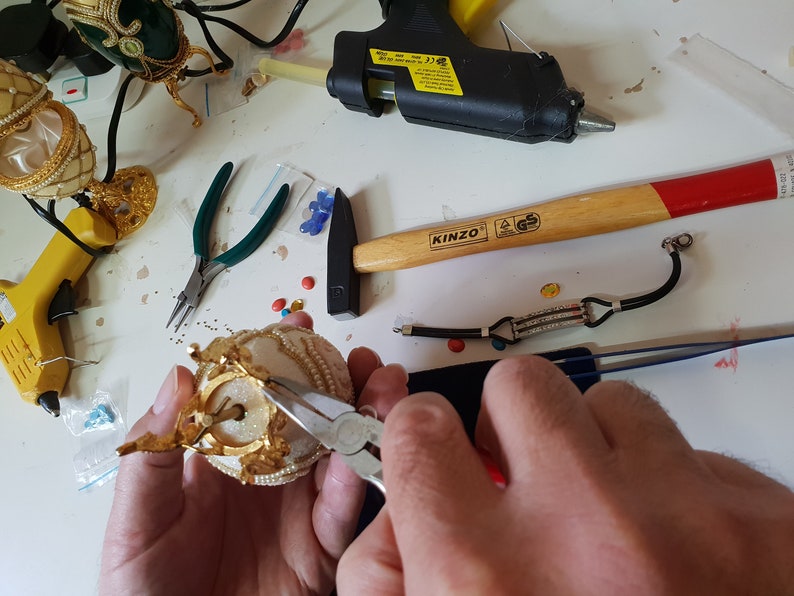 May include: A person is using pliers to attach a gold-colored crown to a white and gold decorative egg. The egg is sitting on a white surface. There are various tools and craft supplies scattered around the egg, including a hammer, a glue gun, and a pair of tweezers.