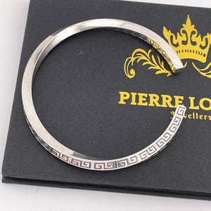 May include: Silver bangle bracelet with a Greek key design. The bracelet is open and has a slightly curved shape. The bracelet is on a black background with the text "PIERRE LO" and "Jewellers" visible.