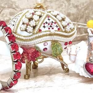 May include: A red jeweled bracelet with a silver band and a white porcelain egg-shaped trinket box with red and green jeweled accents. A silver chain necklace with a red jeweled heart pendant is also pictured.