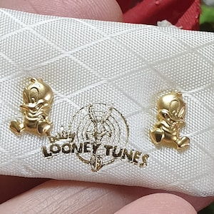 May include: A pair of gold-colored earrings featuring a cartoon duck character. The earrings are in a white velvet pouch with the words "Looney Tunes" printed on it.