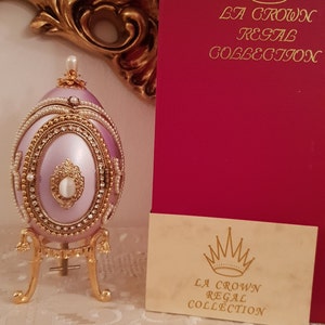 May include: A delicate, hand-painted porcelain egg with a lavender finish, adorned with gold trim, pearls, and rhinestones. It sits on a gold stand and is part of the "La Crown Regal Collection".