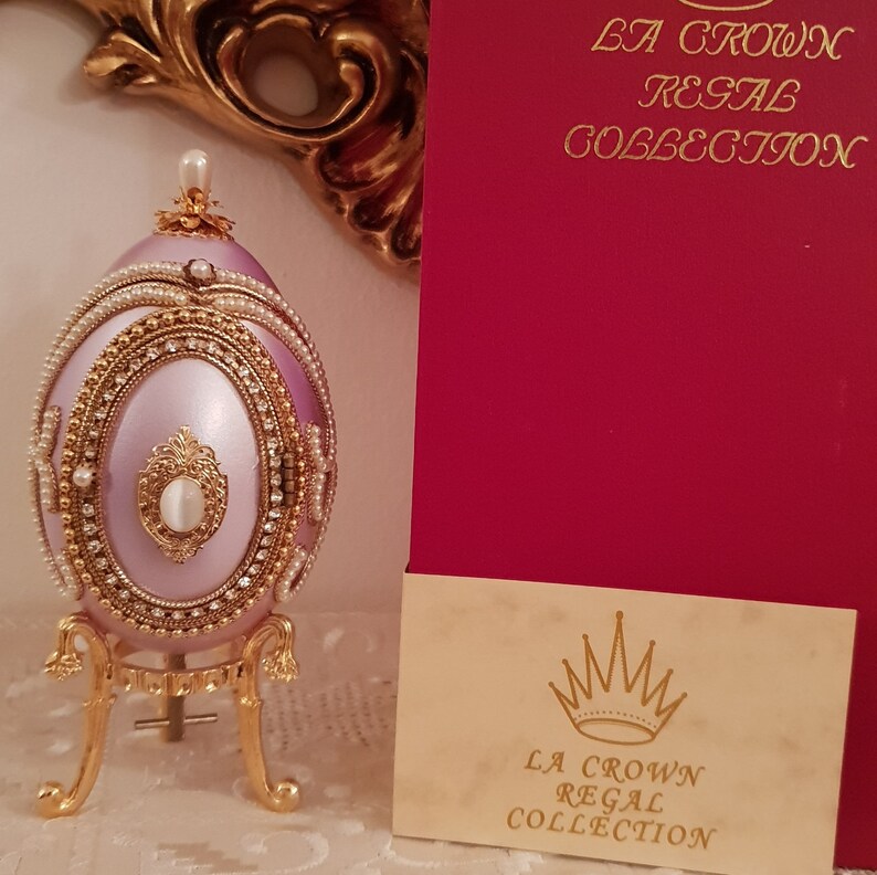 May include: A delicate, hand-painted porcelain egg with a lavender finish, adorned with gold trim, pearls, and rhinestones. It sits on a gold stand and is part of the "La Crown Regal Collection".