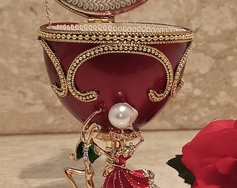 May include: A red and gold egg-shaped trinket box with a hinged lid. The box is decorated with gold beading and a detailed design. The lid is open, revealing a white interior and a small pearl. The box is sitting on a gold base with a sculpted scene of two dancers.