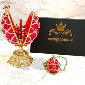 May include: A red and gold jeweled egg with a detailed design opens to reveal a miniature castle inside. The egg is on a gold stand and comes with a matching pendant on a gold chain. The box it comes in is black with gold lettering that reads "Pierre Lorren Jewellers".