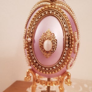 May include: A pink jeweled egg music box with a gold stand. The egg is decorated with pearls and rhinestones. The stand has three legs and a round base.