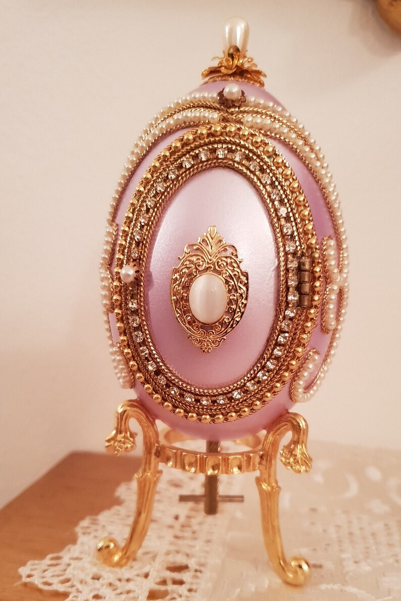 May include: A pink jeweled egg music box with a gold stand. The egg is decorated with pearls and rhinestones. The stand has three legs and a round base.
