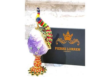 May include: A white and purple egg-shaped trinket box with a gold base and a peacock figurine on top. The peacock is yellow, green, blue, and red. The box is decorated with purple flowers and green leaves. The box is on a white surface. The box is labelled "Pierre Lorren Jewellers".