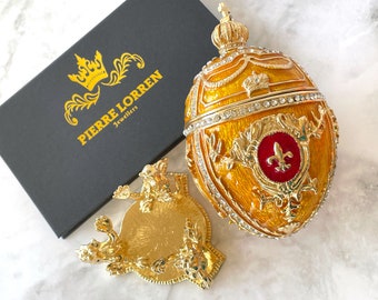 May include: A gold and yellow jeweled egg with a red and gold fleur-de-lis design. The egg is on a gold stand with a crown design. The egg is a decorative piece and is made by Pierre Lorren Jewellers.
