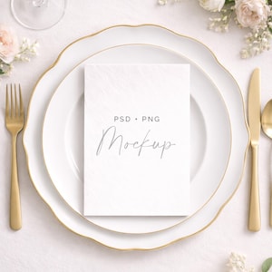 May include: Elegant table setting with white plates, gold cutlery, and a card that says "PSD + PNG Mockup". The plates have a gold trim. A glass of wine and floral arrangements complete the scene.