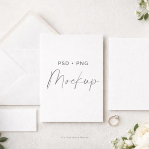 May include: A styled mockup with white stationery, including an envelope, cards, and a business card, alongside white flowers and a gold ring. The central card features the word "Mockup" in script, with "PSD + PNG" above it.