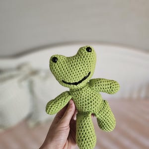 May include: A handmade, light green crochet frog toy with black eyes and a smiling mouth. The frog has small arms and legs, and is held up in front of a soft, blurred background. This plush toy is perfect for a child.