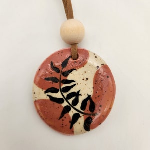 May include: A round ceramic pendant with a leaf design. The pendant features a reddish-brown and cream color scheme with black leaf details. It hangs from a brown leather cord and a wooden bead.