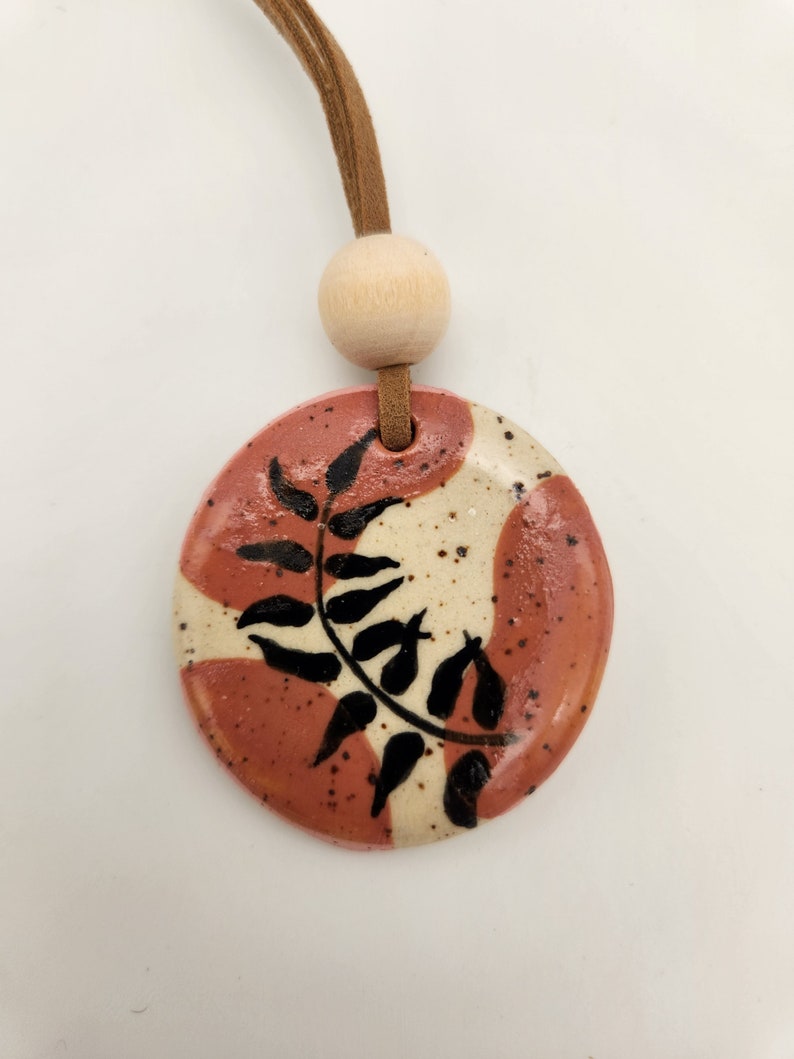 May include: A round ceramic pendant with a leaf design. The pendant features a reddish-brown and cream color scheme with black leaf details. It hangs from a brown leather cord and a wooden bead.
