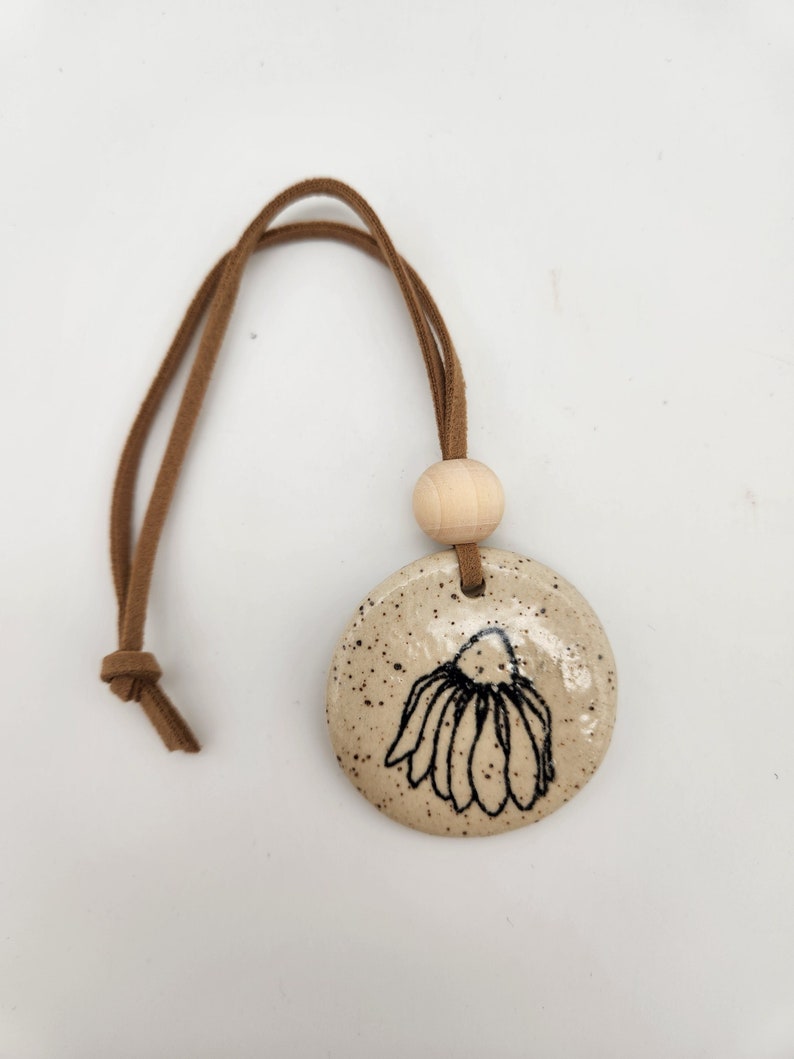 K&ouml;nnte beinhalten: Runder, gesprenkelter Keramikanh&auml;nger mit handgezeichnetem schwarzem Blumendesign. Er hat eine braune Wildlederschnur und eine Holzperle zum Aufh&auml;ngen. Der Anh&auml;nger hat eine hellbeige Farbe.