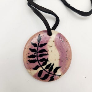 May include: A round pendant necklace with a black leather cord. The pendant is ceramic, featuring a design of a black leaf against a background of cream and lavender. The pendant is approximately 1.5 inches in diameter.