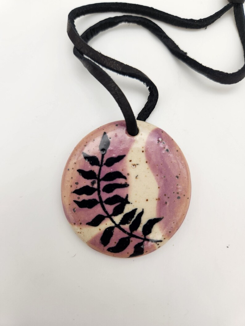 May include: A round pendant necklace with a black leather cord. The pendant is ceramic, featuring a design of a black leaf against a background of cream and lavender. The pendant is approximately 1.5 inches in diameter.