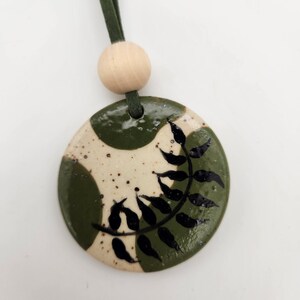 May include: A handmade ceramic pendant with a green and cream speckled design. The pendant features a black leaf pattern and is strung on a green suede cord with a wooden bead. This unique necklace is a perfect accessory.