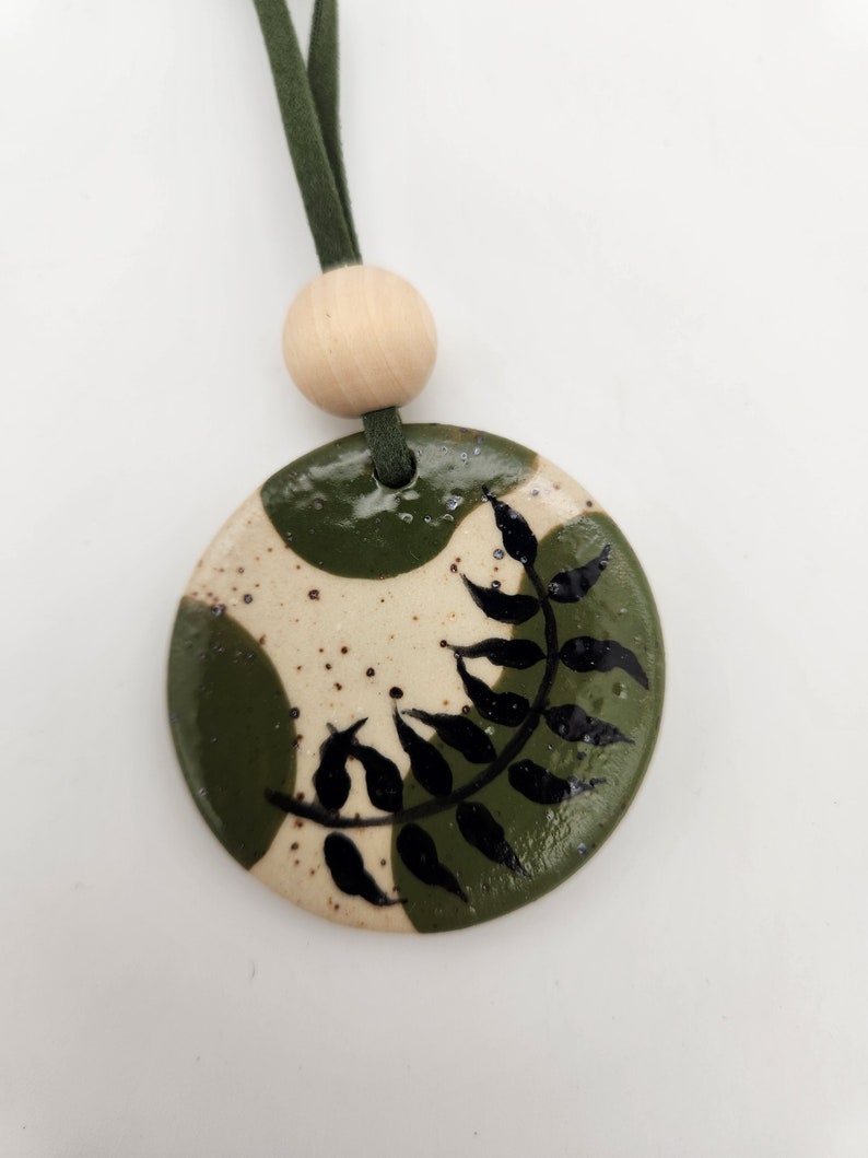 May include: A handmade ceramic pendant with a green and cream speckled design. The pendant features a black leaf pattern and is strung on a green suede cord with a wooden bead. This unique necklace is a perfect accessory.