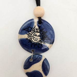 Op de afbeelding: Een keramische hangerketting met twee blauwe en witte cirkelvormige hangers. De bovenste hanger heeft een zon- en maanontwerp. De ketting heeft een zwart koord en een houten kraal.
