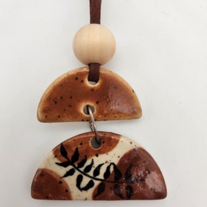 May include: A ceramic pendant with a brown leather cord. The pendant has two half-circle shapes in brown and cream with a leaf design. A wooden bead is above the ceramic pieces. The pendant is a decorative item.