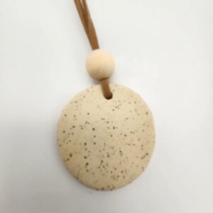 May include: A round, speckled, beige ceramic pendant with a small hole at the top. It is attached to a light brown suede cord and a wooden bead. The pendant is a neutral color and has a rustic, handmade appearance.