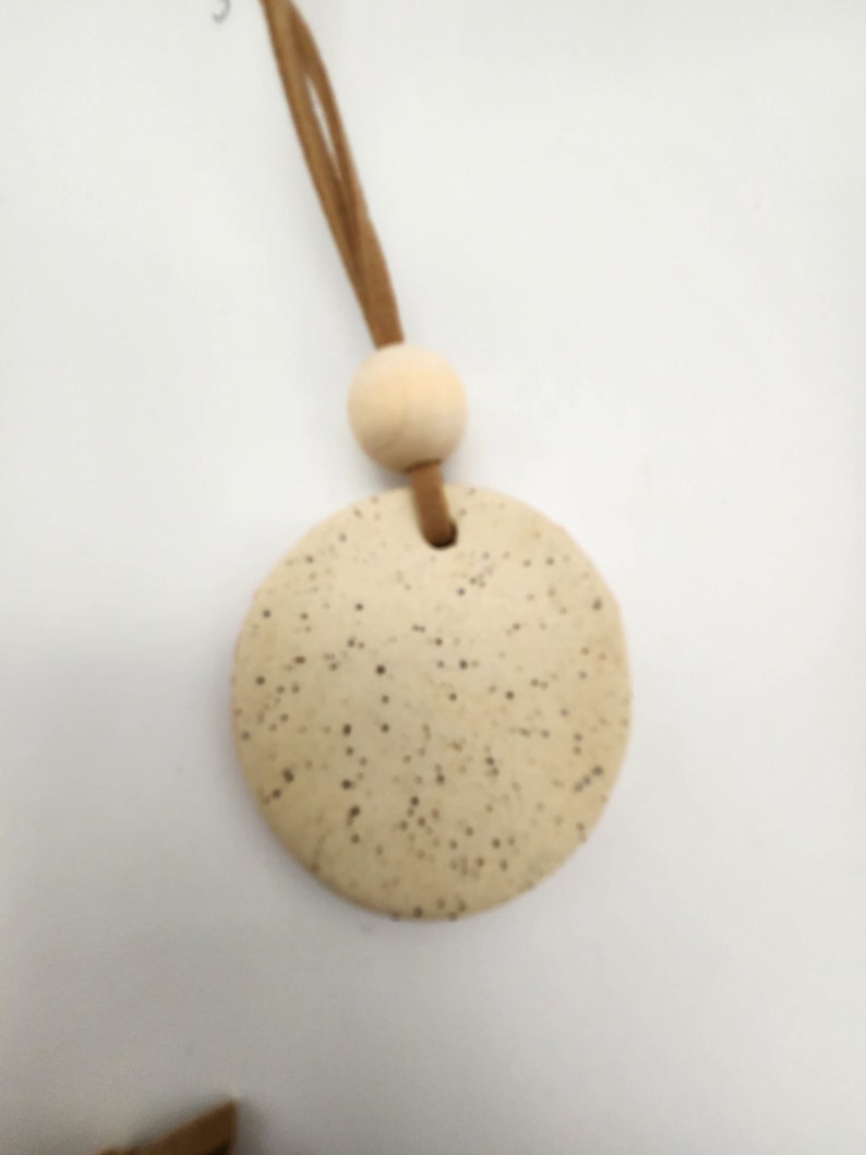 May include: A round, speckled, beige ceramic pendant with a small hole at the top. It is attached to a light brown suede cord and a wooden bead. The pendant is a neutral color and has a rustic, handmade appearance.