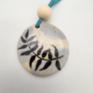 May include: A ceramic pendant with a hand-painted black leaf design. The pendant is round, with a light blue suede cord and a wooden bead. The background of the pendant is a mix of beige and gray.