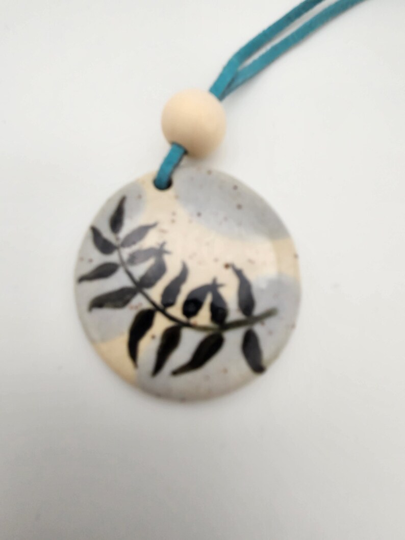 May include: A ceramic pendant with a hand-painted black leaf design. The pendant is round, with a light blue suede cord and a wooden bead. The background of the pendant is a mix of beige and gray.