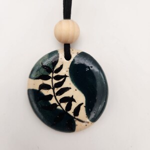 May include: A ceramic pendant necklace with a black cord. The pendant is a rounded oval shape with a cream-colored background and dark teal and black accents. A black fern design is painted on the pendant. A wooden bead is above the pendant.
