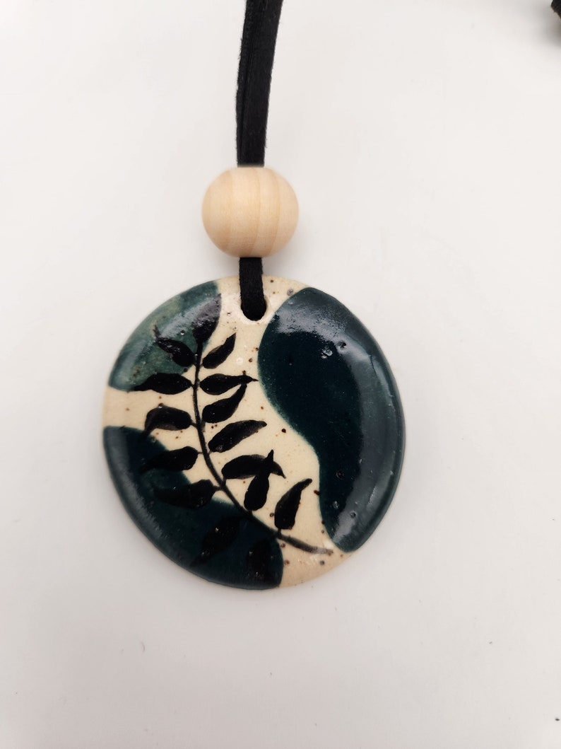 May include: A ceramic pendant necklace with a black cord. The pendant is a rounded oval shape with a cream-colored background and dark teal and black accents. A black fern design is painted on the pendant. A wooden bead is above the pendant.