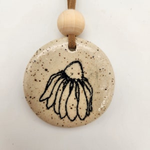 K&ouml;nnte beinhalten: Ein runder, gesprenkelter Keramikanh&auml;nger mit einem schwarzen Blumendesign. Der Anh&auml;nger ist an einer braunen Wildlederschnur und einer Holzperle befestigt. Der Anh&auml;nger ist beige mit dunkelbraunen Sprenkeln.