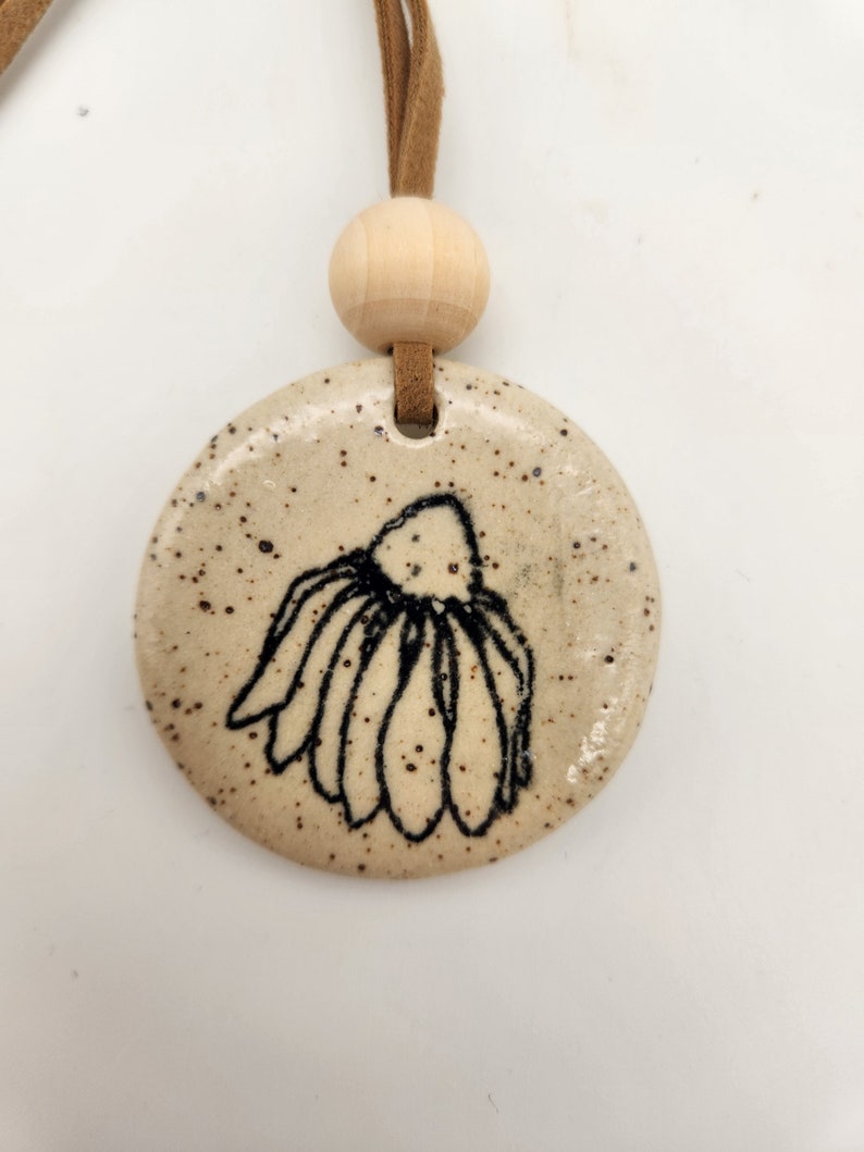 K&ouml;nnte beinhalten: Ein runder, gesprenkelter Keramikanh&auml;nger mit einem schwarzen Blumendesign. Der Anh&auml;nger ist an einer braunen Wildlederschnur und einer Holzperle befestigt. Der Anh&auml;nger ist beige mit dunkelbraunen Sprenkeln.