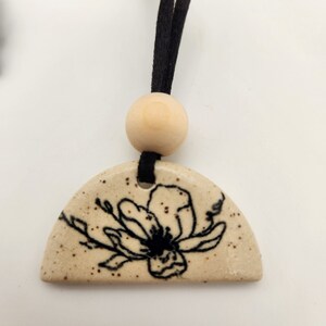 Könnte beinhalten: Beigefarbener Keramikanhänger mit schwarzem Blumenmuster, aufgehängt an einer schwarzen Kordel und einer Holzperle. Der Anhänger ist halbkreisförmig mit einer gesprenkelten Textur, und die Blume ist handgezeichnet.
