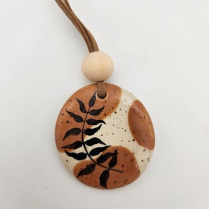 May include: A round ceramic pendant with a brown and cream speckled design, featuring a black leaf pattern. It hangs from a brown cord with a wooden bead. The pendant has a natural, earthy aesthetic.