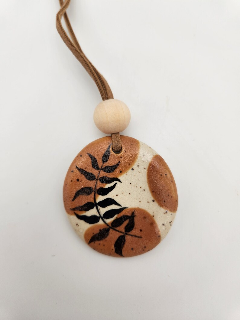 May include: A round ceramic pendant with a brown and cream speckled design, featuring a black leaf pattern. It hangs from a brown cord with a wooden bead. The pendant has a natural, earthy aesthetic.