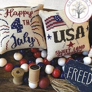 Könnte beinhalten: Zwei kleine Kissenbezüge mit Kreuzstich-Stickerei. Ein Kissenbezug zeigt eine rot-weiß-blaue amerikanische Flagge mit dem Text "USA" und "Sweet Land of Liberty". Der andere Kissenbezug zeigt den Text "Happy 4th of July" mit rot-weiß-blauen Feuerwerkskörpern.