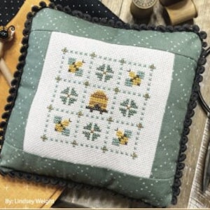 Puede incluir: Una almohada de punto de cruz verde y blanca con un diseño de colmena y abejas. La almohada está adornada con flecos negros y tiene el texto "August Nine Patch" y "Primrose Cottage Stitches".