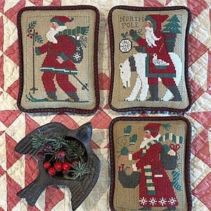 May include: Four decorative Christmas pillows with cross-stitch designs of Santa Claus in red and white outfits, and a black bird-shaped container holding greenery and red berries. The pillows have a brown border and are set against a red and white quilt.